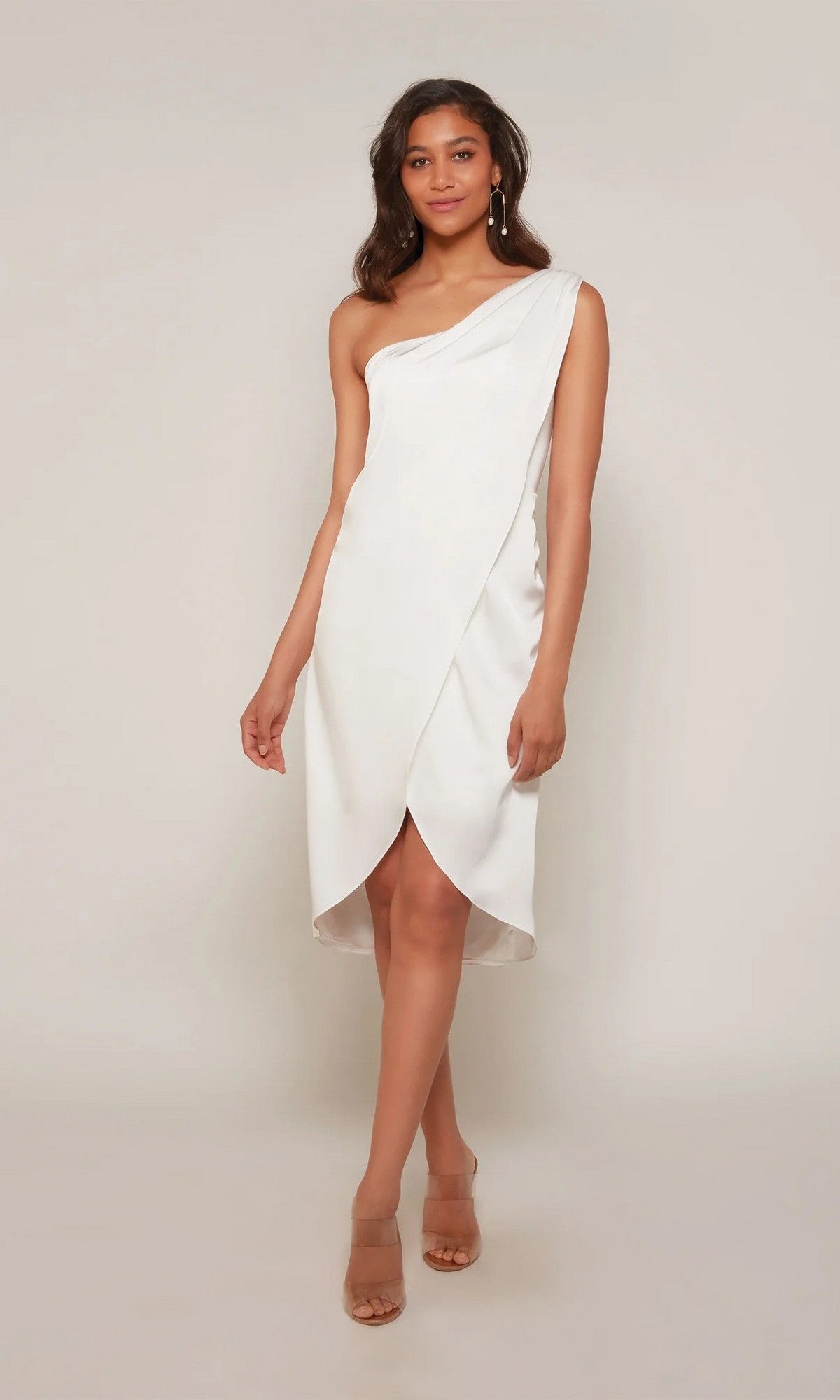 A woman stands in the Alyce 70038 knee-length white party dress with a one-shoulder design and tulip hem, paired with nude heels and drop earrings against a plain background.